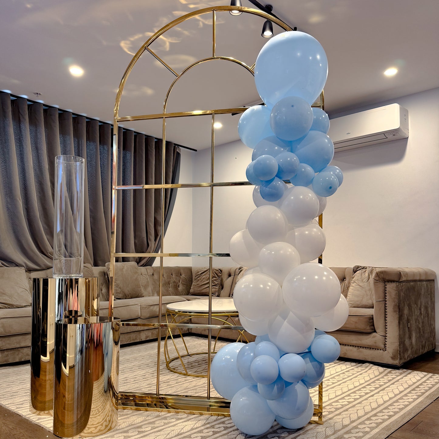 A metal arch backdrop decorated with a series of round balloons in shades of blue, gold, and white, set in a room with a grey sofa and a gold-colored arch.