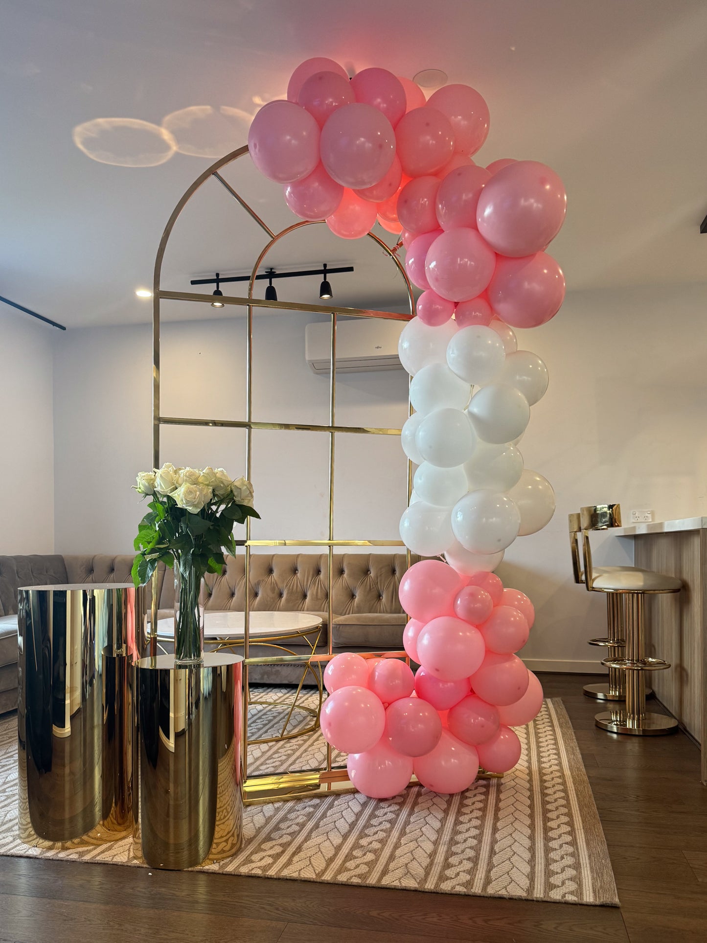 Gold metal arch decorated with pink and white balloon garland, next to gold cylindrical stands holding a vase of white roses in a modern living room