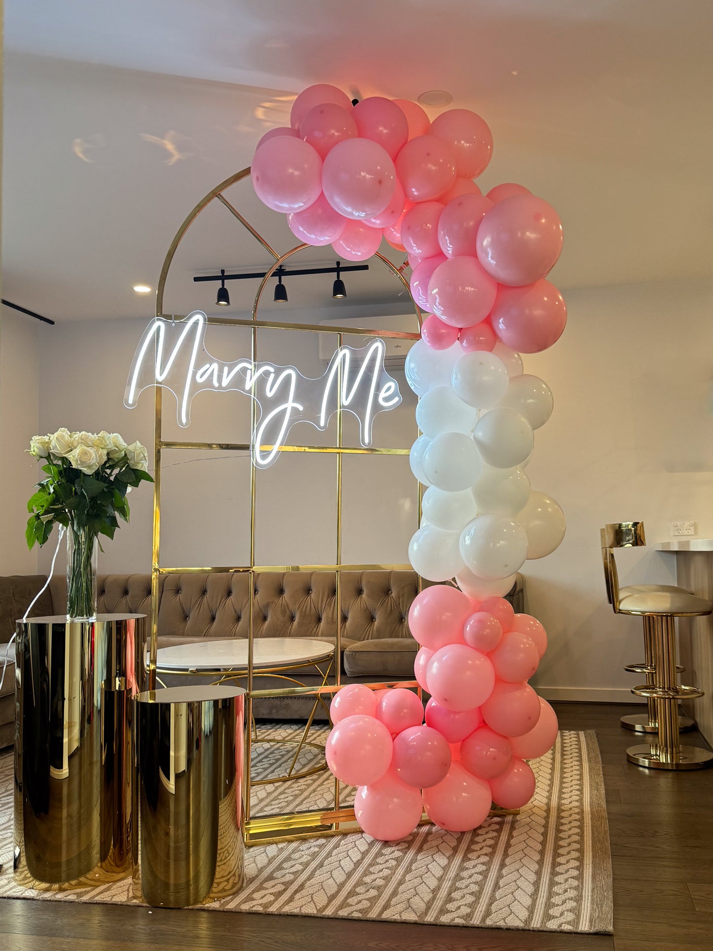 Gold metal arch with a white neon ‘Marry Me’ sign, decorated with clusters of pink and white balloons, flanked by gold plinths holding a vase of white roses in a modern living room
