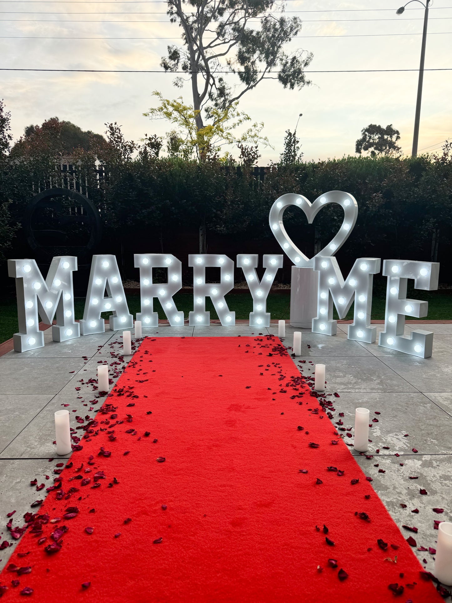 Outdoor proposal setup with illuminated letters spelling 'MARRY ME' and a heart above, a red carpet lined with candles and scattered rose petals