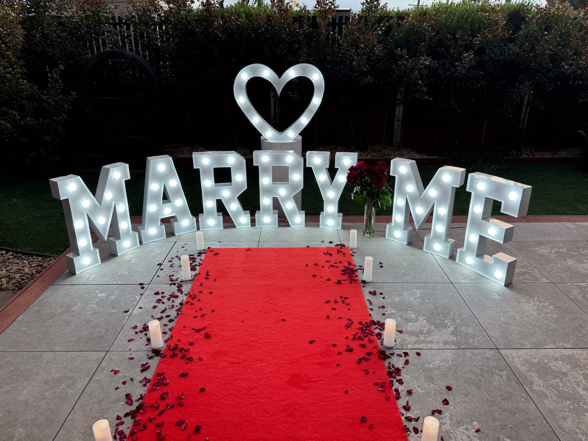 Romantic outdoor setup featuring illuminated marquee letters spelling ‘MARRY ME’ with a heart symbol, a vase of red roses, candlelit pathway, and scattered rose petals on a red carpet