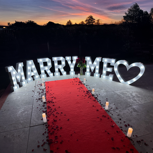 A proposal setup with 'MARRY ME' LED lettering, a heart shape, a red carpet, candles, and rose petals.