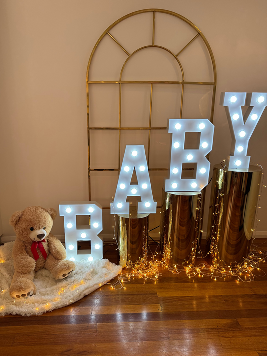 Decorative setup with 'BABY' light-up letters, teddy bear, and fairy lights on a wooden floor.