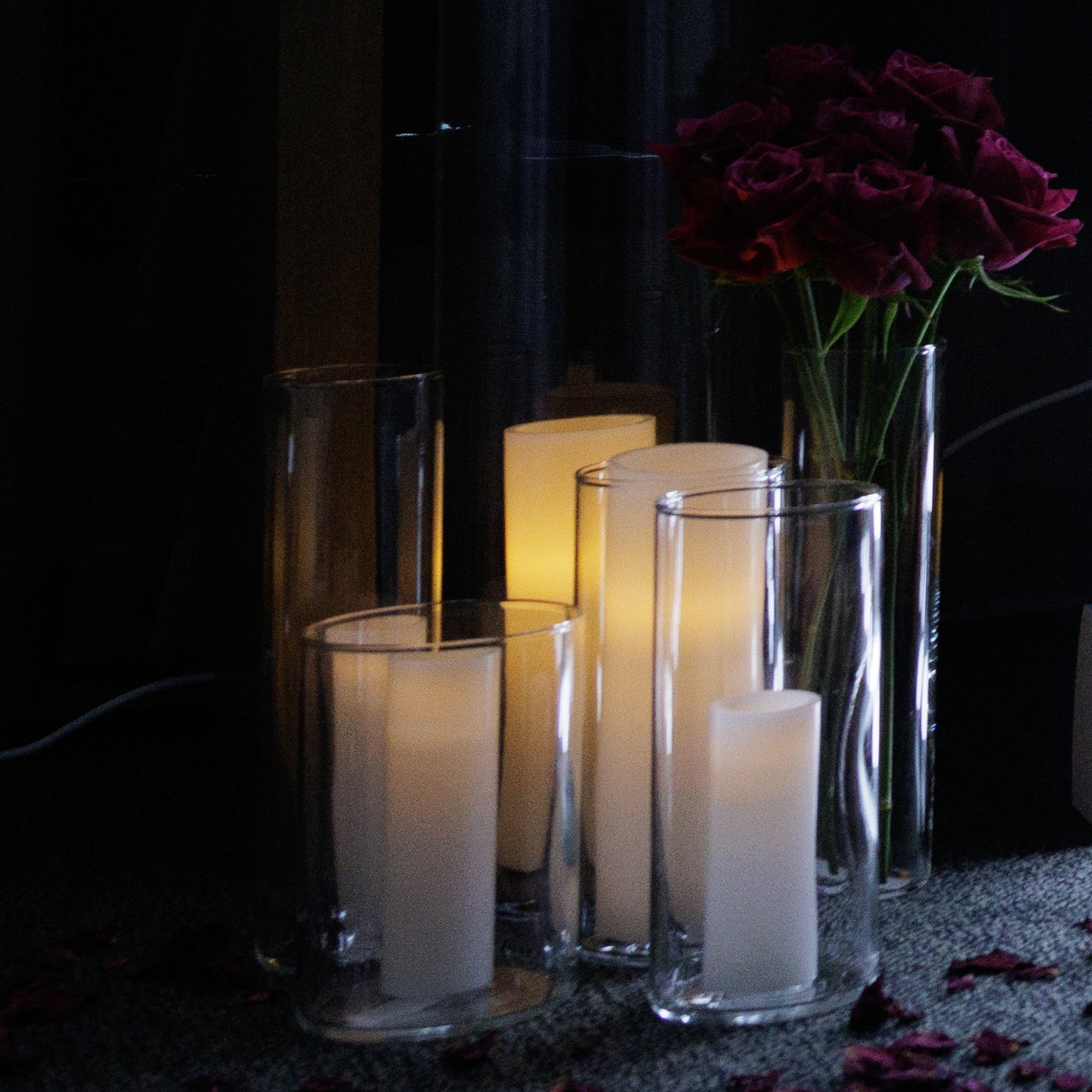 Cluster of lit candles in tall glass holders next to a vase of deep red roses, placed on a dark carpet scattered with rose petals.