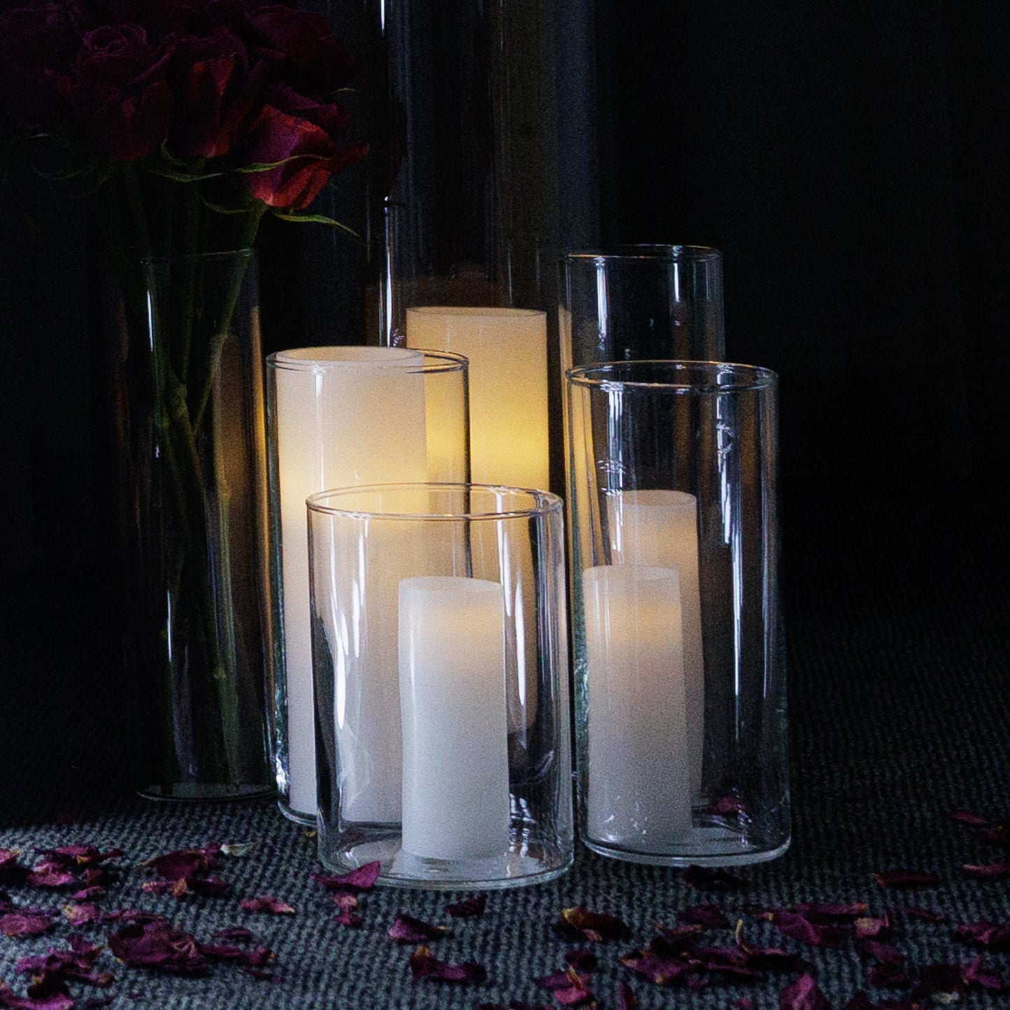 A set of clear glass vases of varying heights arranged on a surface, with lit candles inside the middle two vases, and scattered rose petals around the arrangement.