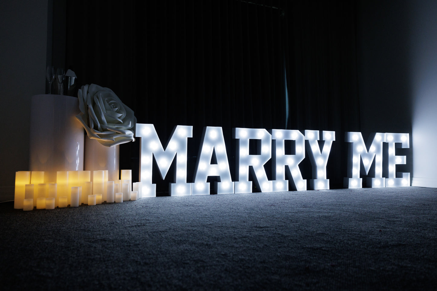 Illuminated 'Marry Me' marquee letters with LED candles and large white rose decoration on a carpeted floor, set for a romantic proposal