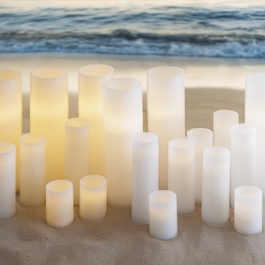 A collection of white, cylindrical electronic candles arranged on a sandy surface with a blurred background featuring waves and sky.