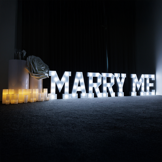 An 'Marry Me' LED lettering sign with electronic candles, plinths, and rose petals on a dark background.