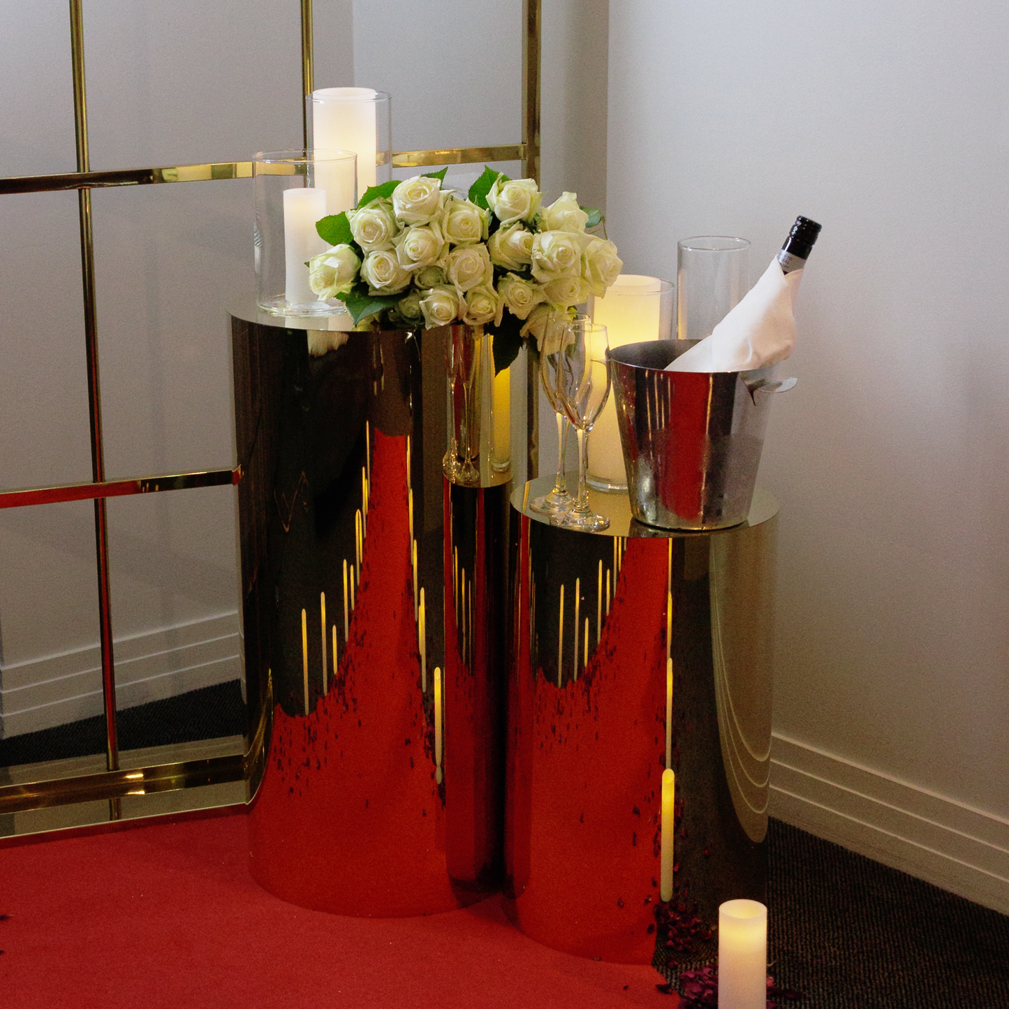 pair of gold plinths on a red carpet, with a bouquet of white roses, large LED candles and a champagne bucket on top