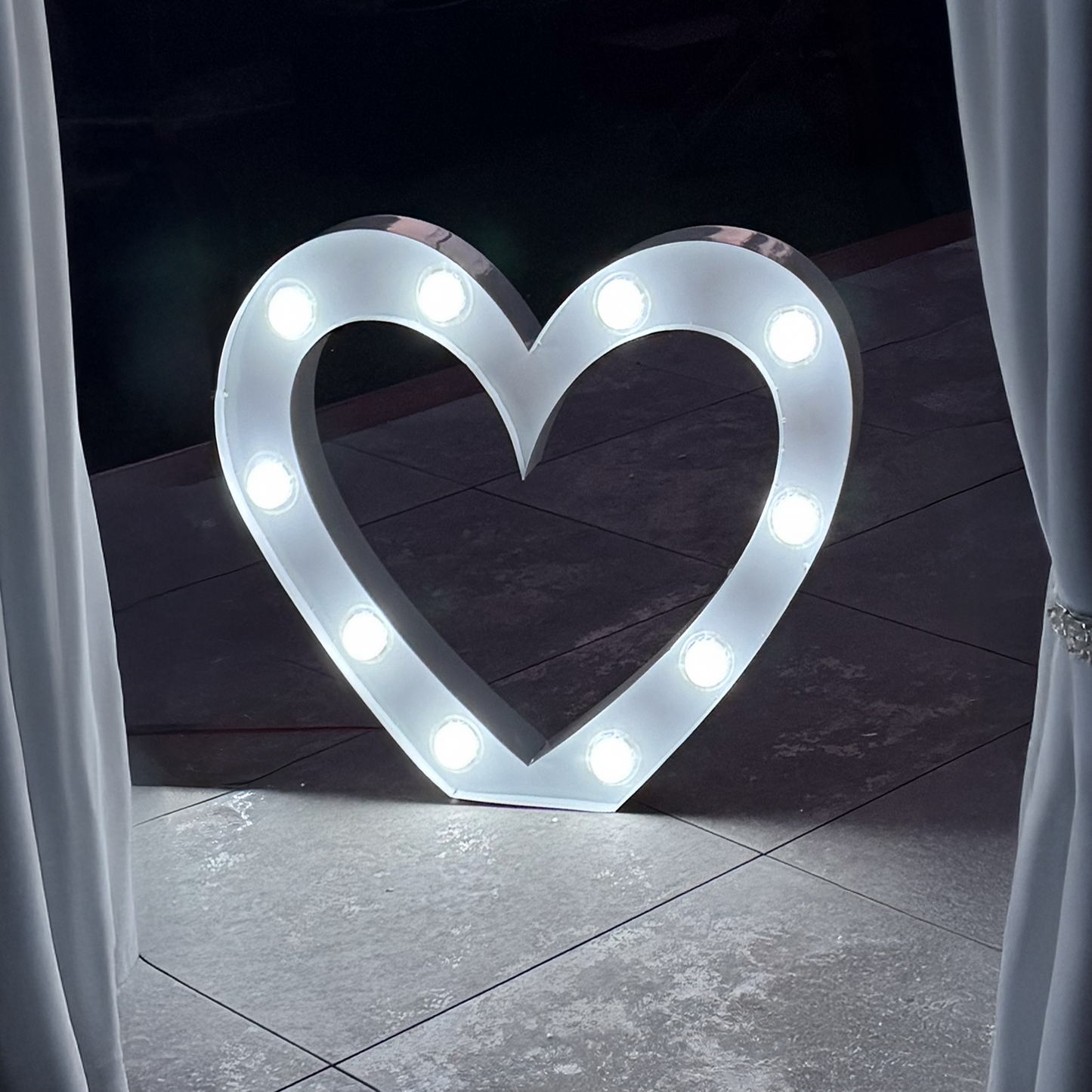 a marquee style light in the shape of a heart, resting on tiles outside at night and framed by curtains
