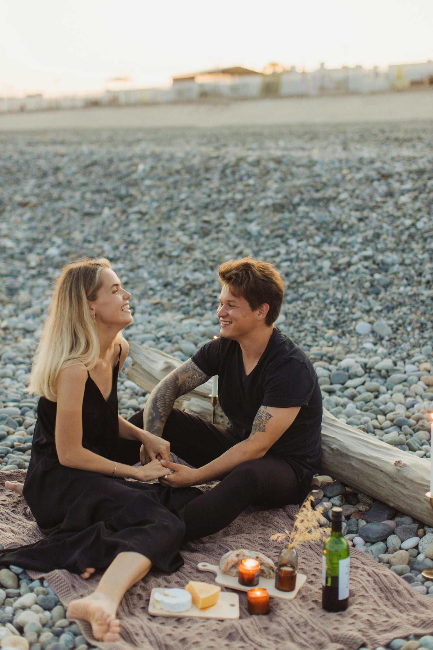 Couple sitting on a blanket by the beach with candles and drinks.