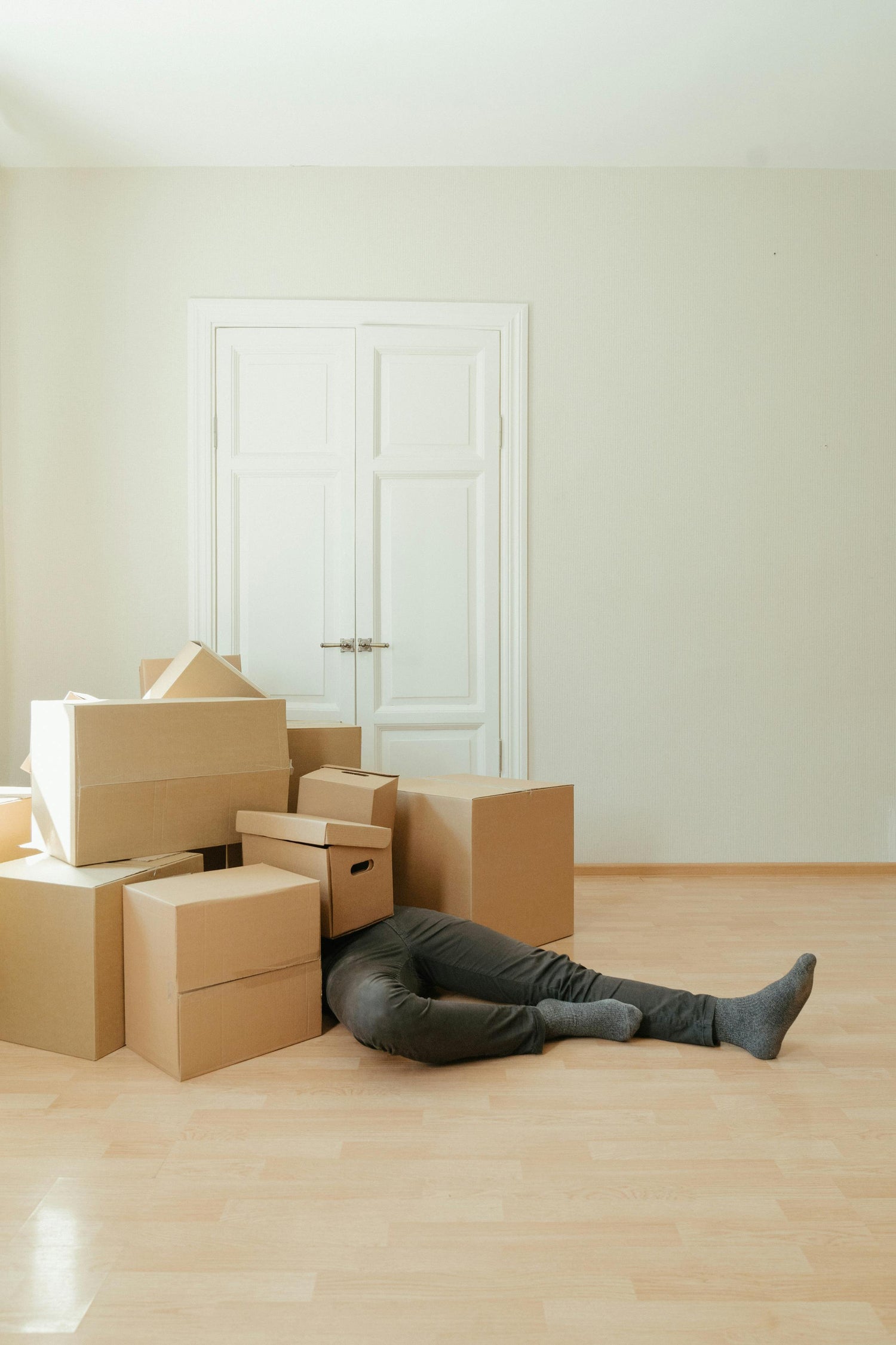 Person lying on a wooden floor surrounded by cardboard boxes in an empty room.
