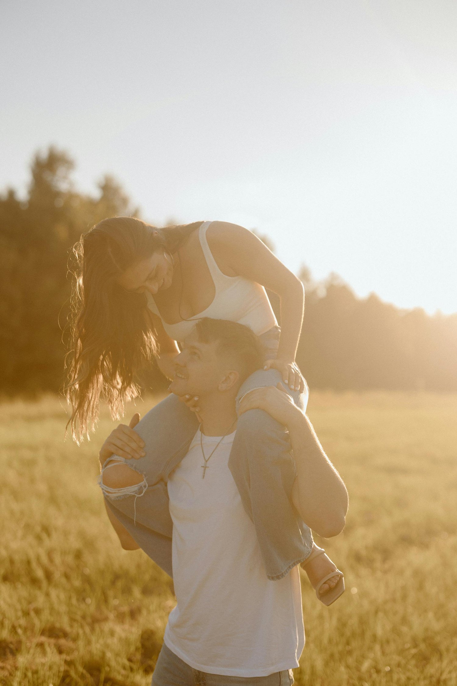 Couple in a sunny field. The woman sits on the mans shoulders as they smile at each other.