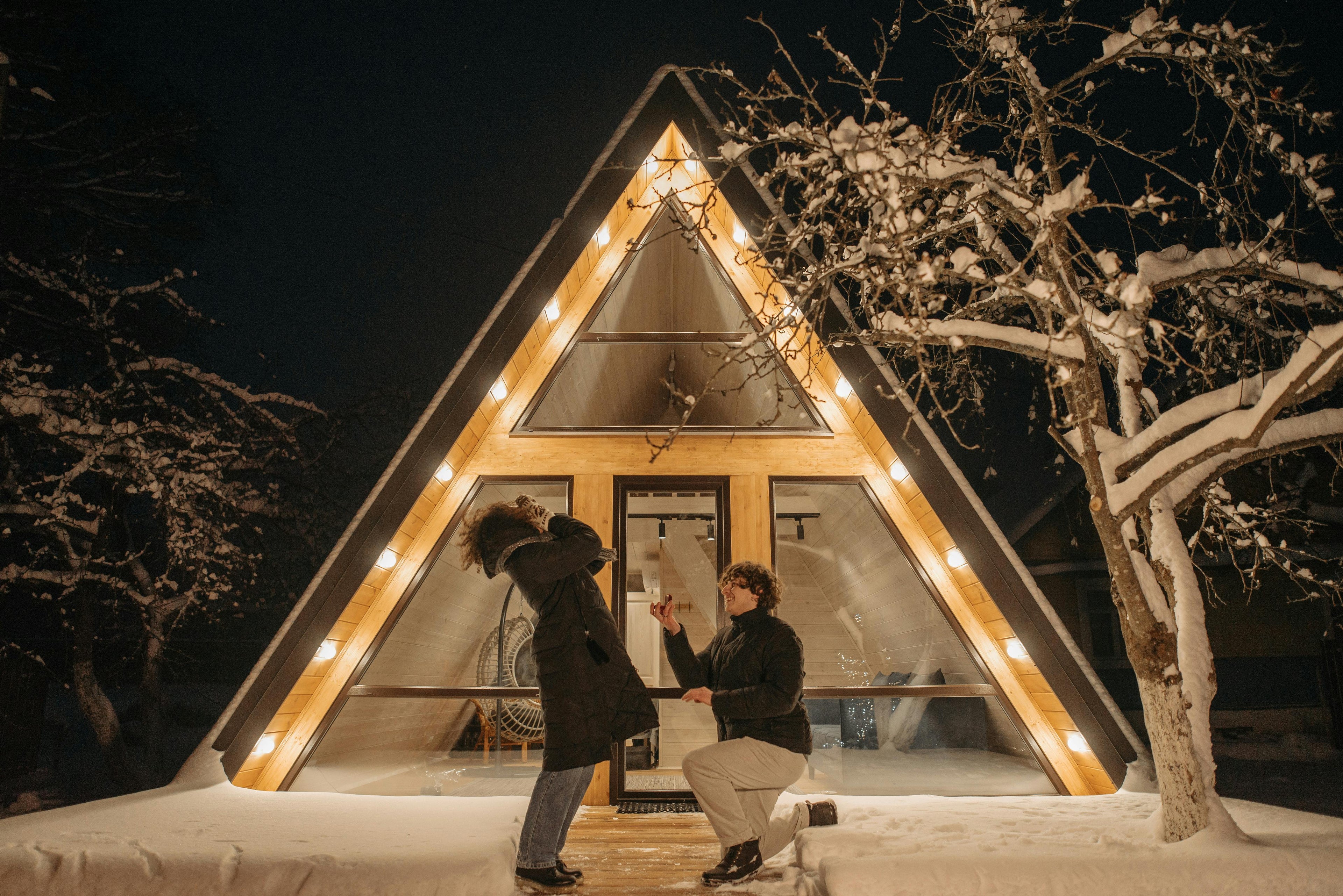Man proposing in front of a triangular glass cabin with lights at night, surrounded by snow.