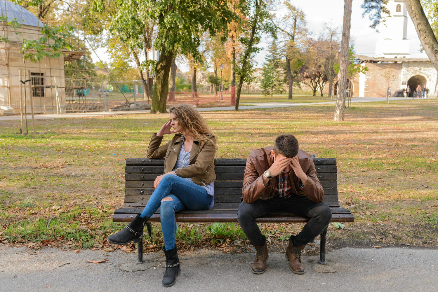 Two people sitting on a bench in a park, one covering their face, indicating distress.