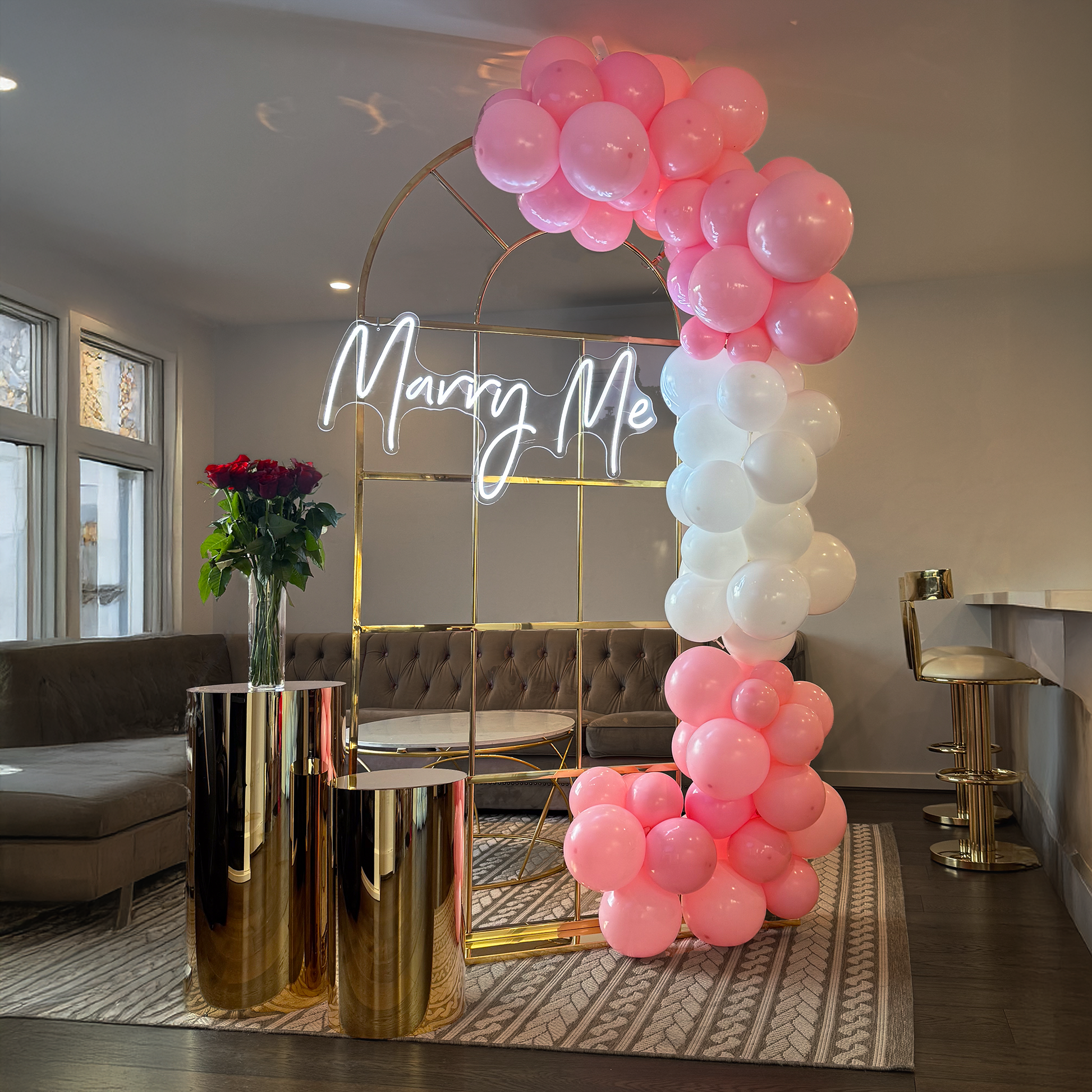 A sophisticated proposal setup with a 'Marry Me' LED sign, gold arch, pink and white balloons, and a floral arrangement in a large vase.