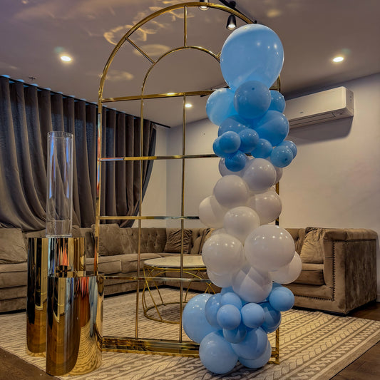 Indoor decorative setup featuring a gold metal arch adorned with clusters of blue and white balloons, positioned beside gold cylindrical stands in a modern living room with neutral-toned sofas and curtains.
