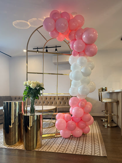 Gold metal arch decorated with pink and white balloon garland, next to gold cylindrical stands holding a vase of white roses in a modern living room