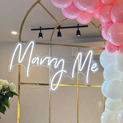 Close-up of a gold metal arch with a white neon ‘Marry Me’ sign, decorated with pink and white balloons and a vase of white roses on the side