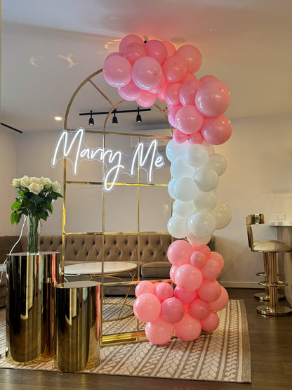 Gold metal arch with a white neon ‘Marry Me’ sign, decorated with clusters of pink and white balloons, flanked by gold plinths holding a vase of white roses in a modern living room