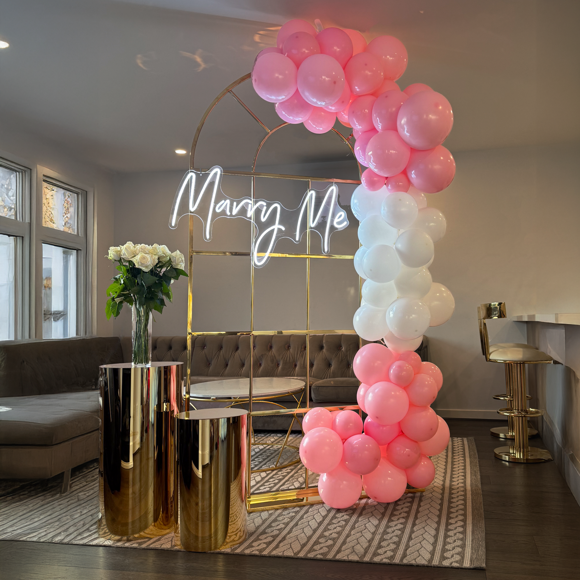 Gold metal arch with a white neon ‘Marry Me’ sign, decorated with clusters of pink and white balloons, flanked by gold plinths holding a vase of white roses in a modern living room