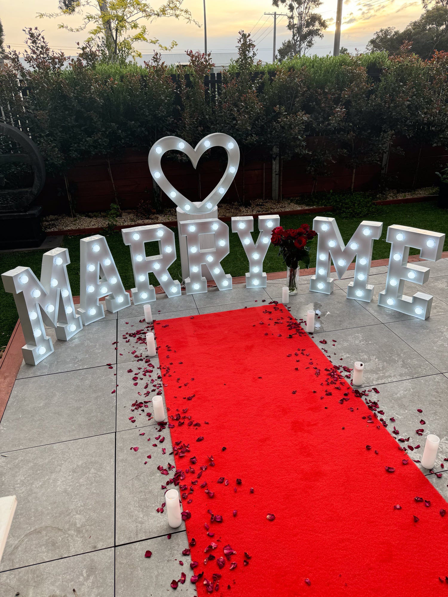 outdoor proposal setup with a red carpet decorated in dried rose petals and surrounded by candles. Marquee light letters spell out "Marry Me" with a matching light up heart. The setup is completed with a bunch of red roses. 