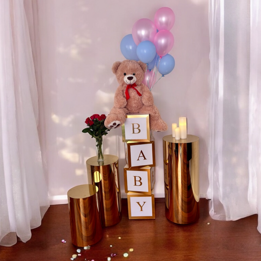 Decorative setup with teddy bear, balloons, and 'BABY' blocks on gold stands against a white curtain background.