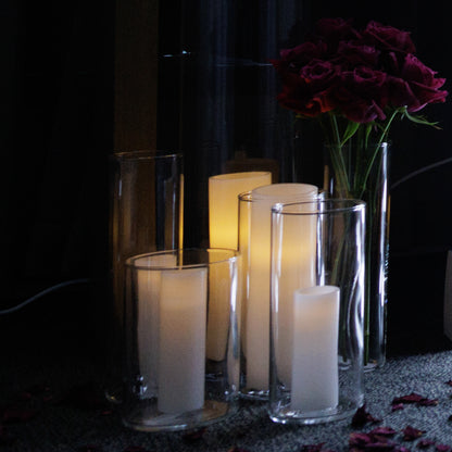 Cluster of lit candles in tall glass holders next to a vase of deep red roses, placed on a dark carpet scattered with rose petals.