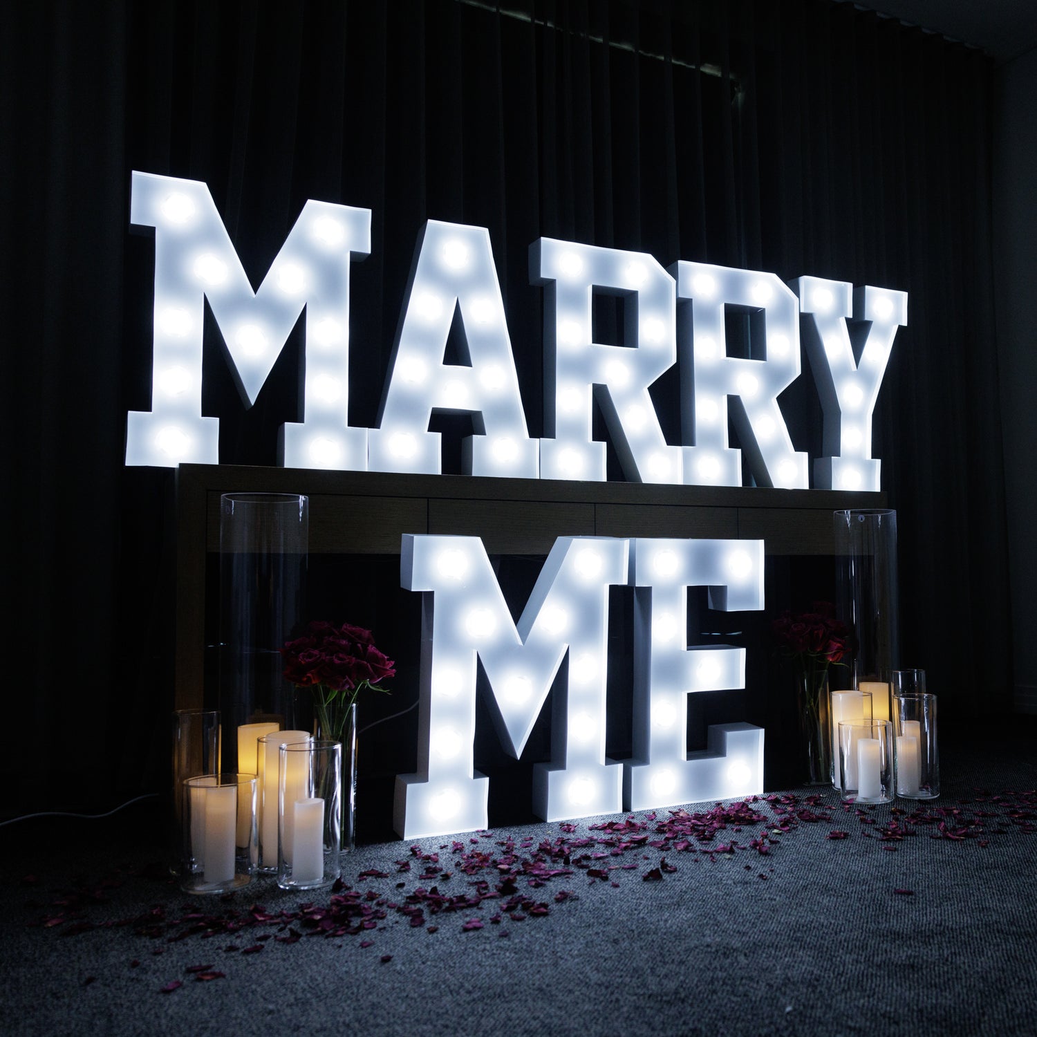 Illuminated marquee light letters spelling out 'MARRY ME' with individual letters on a stand, set against a dark background with candles and rose petals.