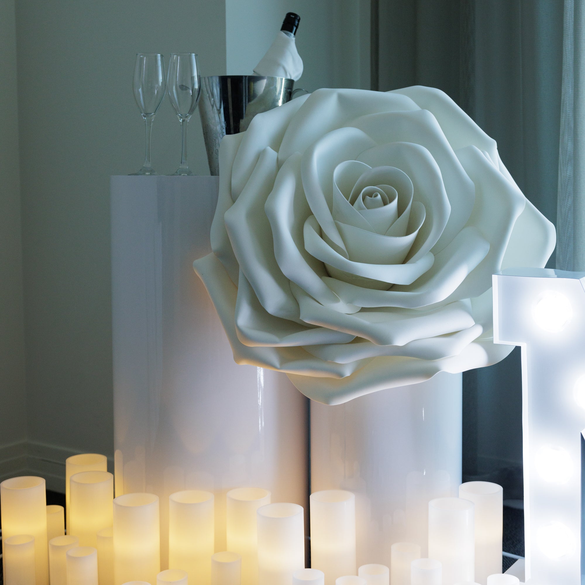 A large artificial rose in white, displayed as a centerpiece on a table with lit candles around it.