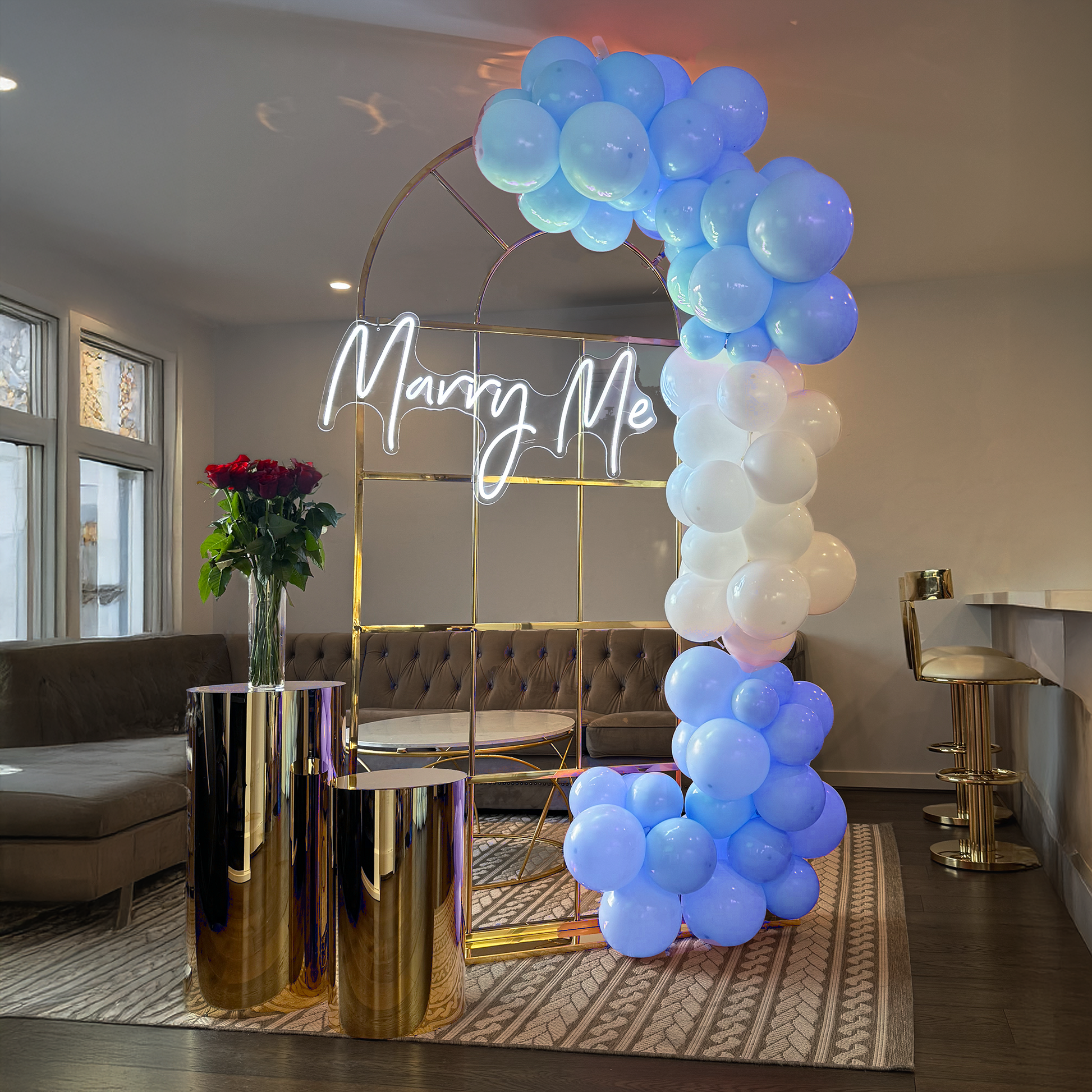 Indoor decorative setup featuring a gold metal arch adorned with a white neon sign spelling out "Marry Me" with clusters of blue and white balloons, positioned beside gold cylindrical stands with a bouquet of red roses in a glass vase.
