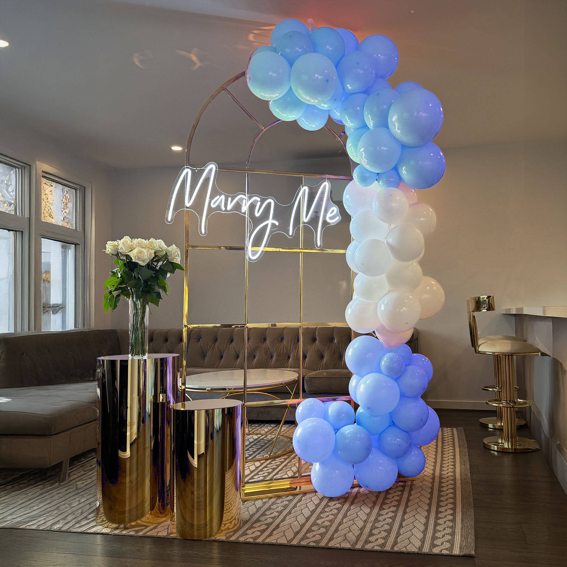 Indoor decorative setup featuring a gold metal arch adorned with a white neon sign spelling out "Marry Me" with clusters of blue and white balloons, positioned beside gold cylindrical stands in a modern living room with neutral-toned sofas and curtains.