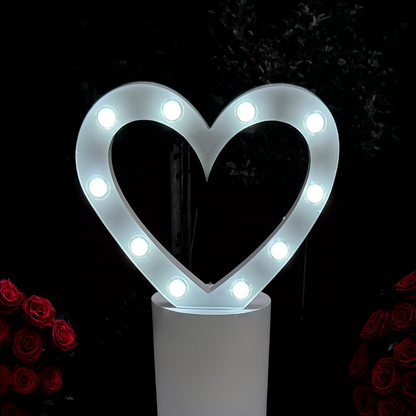 A heart-shaped marquee light symbol displayed at an event, with multiple LED lights illuminated in a warm white color, set against a backdrop of roses and a dark background.