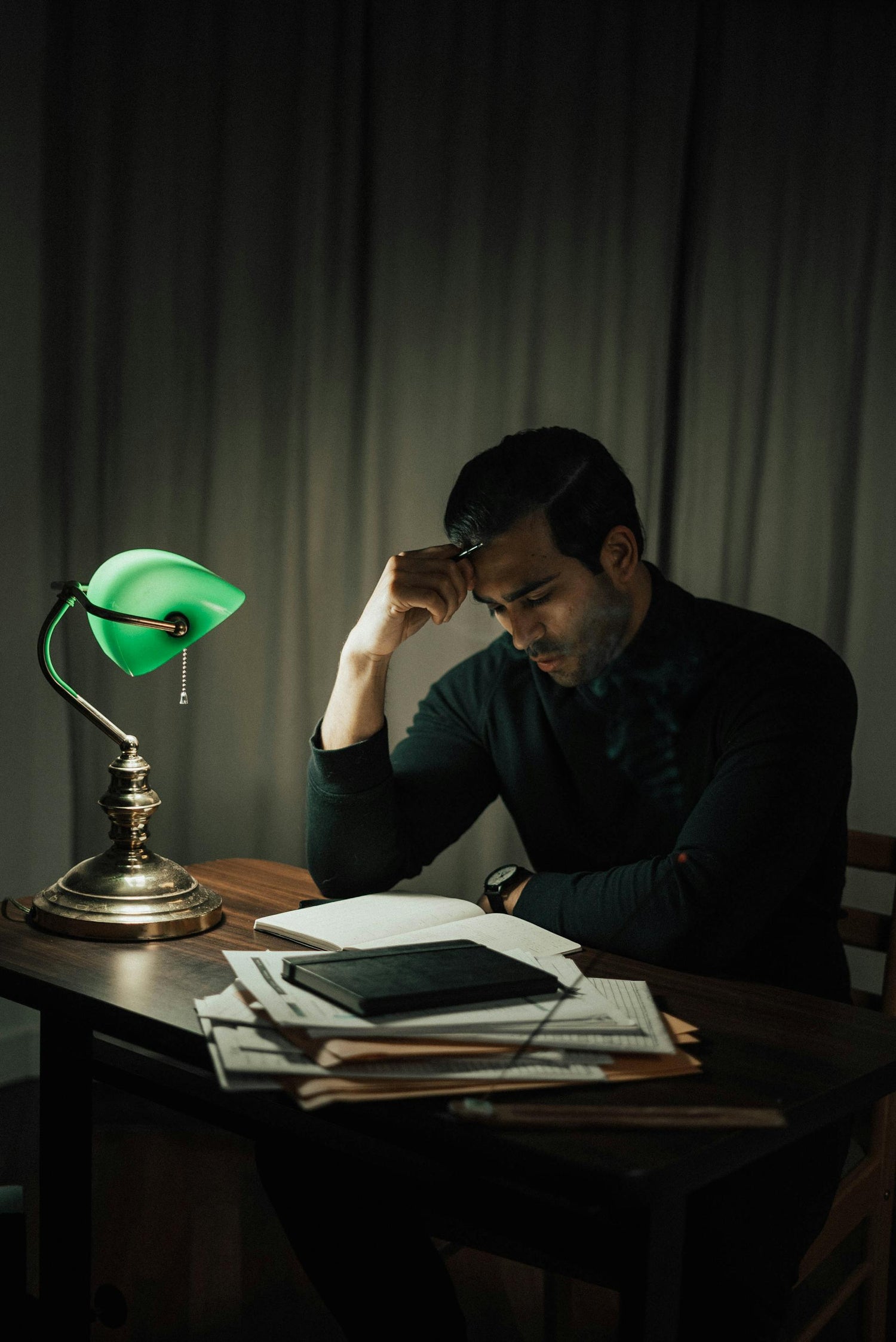 Man sitting at a desk with a green lamp and papers in a dimly lit room